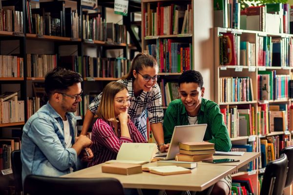 Vier Personen sitzen in einer Bibliothek an einem Tisch und arbeiten gemeinsam mit Laptop und Fachbüchern. Szene zeigt konzentriertes Recherchieren und Prüfen von Informationen.
